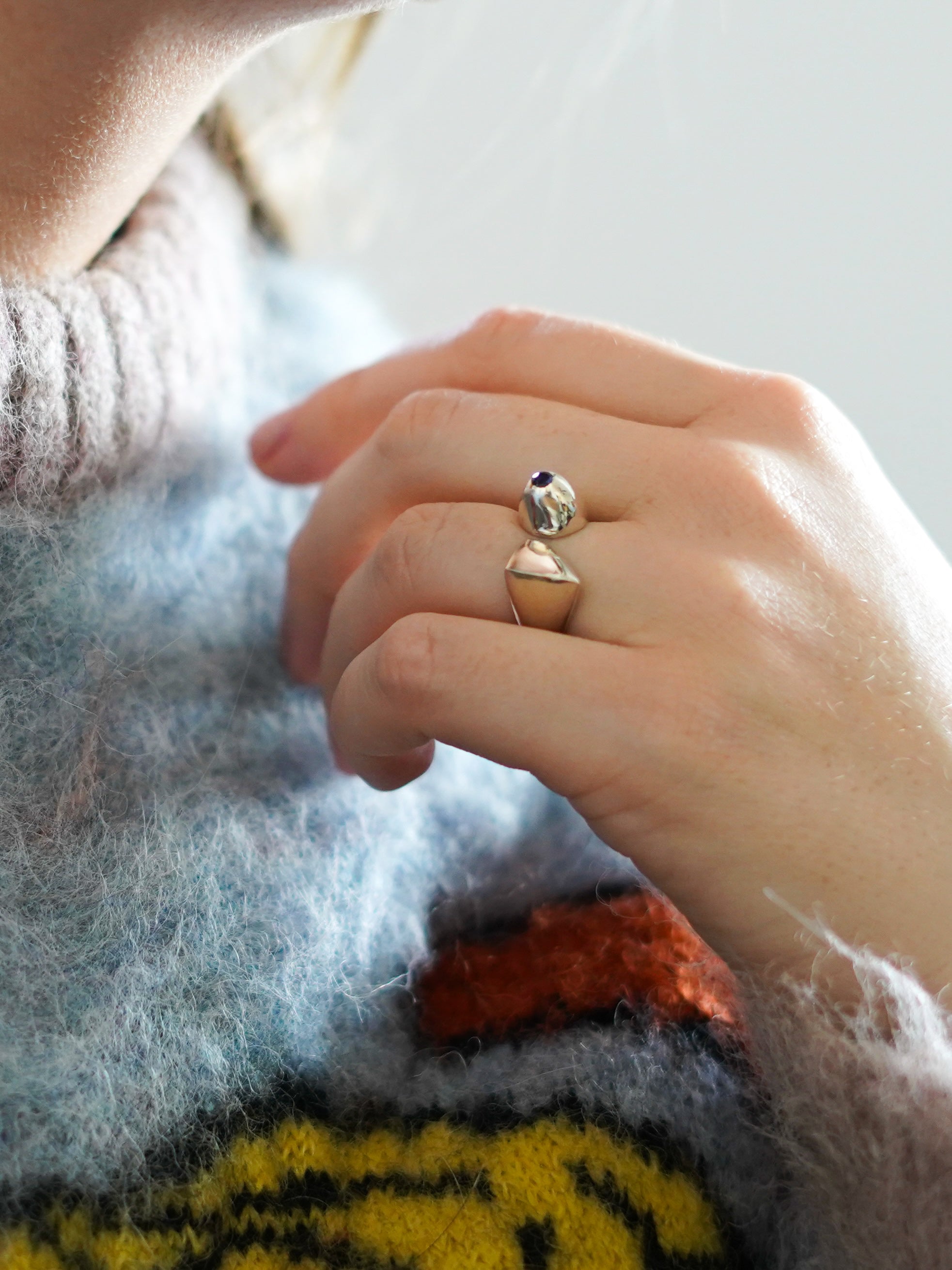 A close-up image of a person's hand resting on a light blue fuzzy sweater.  On their ring finger is a distinctive asymmetrical open silver-colored ring with a small blue violet round gemstone. The ring has a high polish finish and a dome shape on the side where the stone sits.