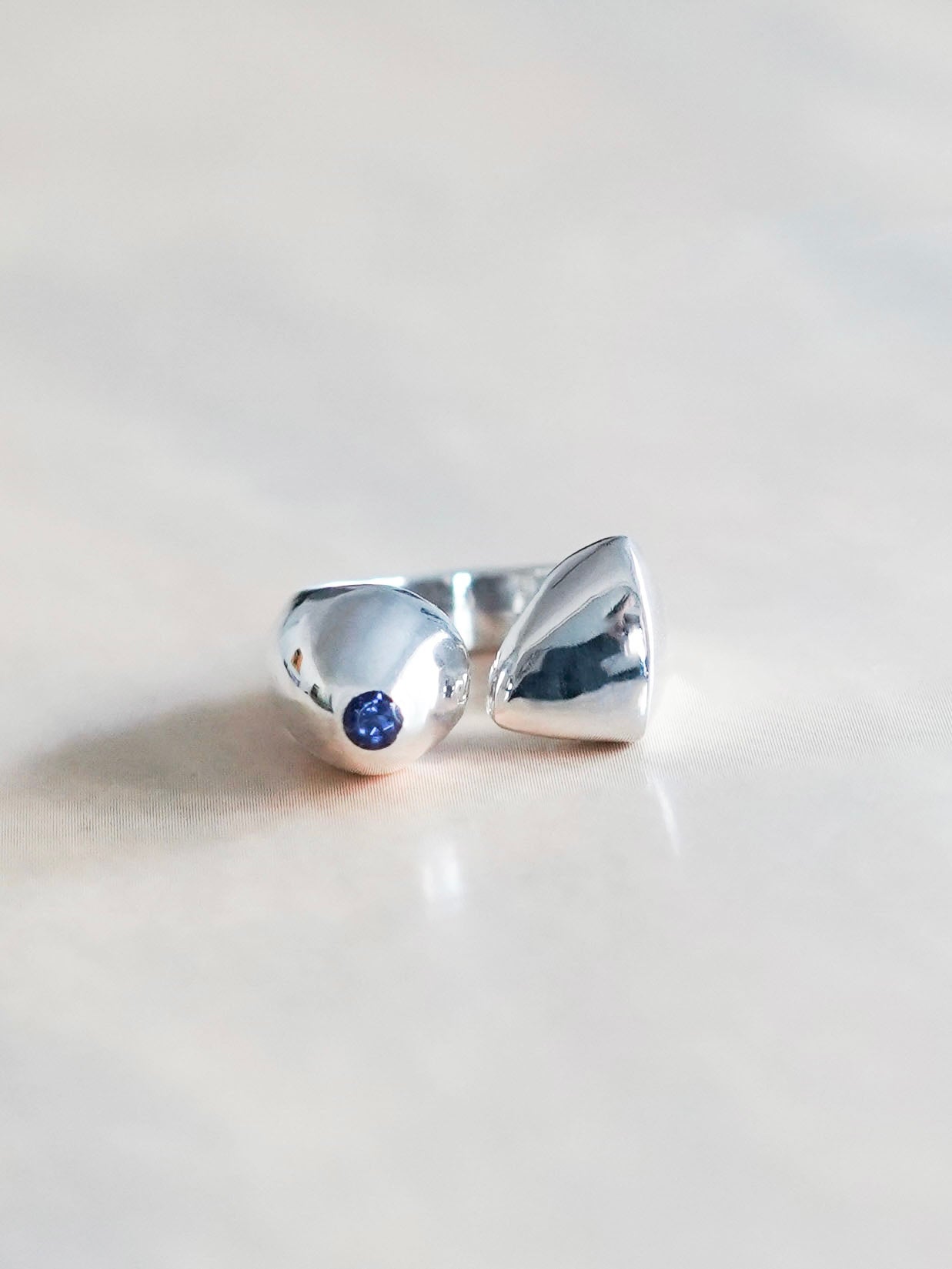 A modern silver ring featuring a round, violet-blue iolite stone set in a smooth, polished bezel. The ring's band splits and curves around the stone, creating an open, asymmetrical design. The ring is photographed on a light neutral background, highlighting its sleek and contemporary style.